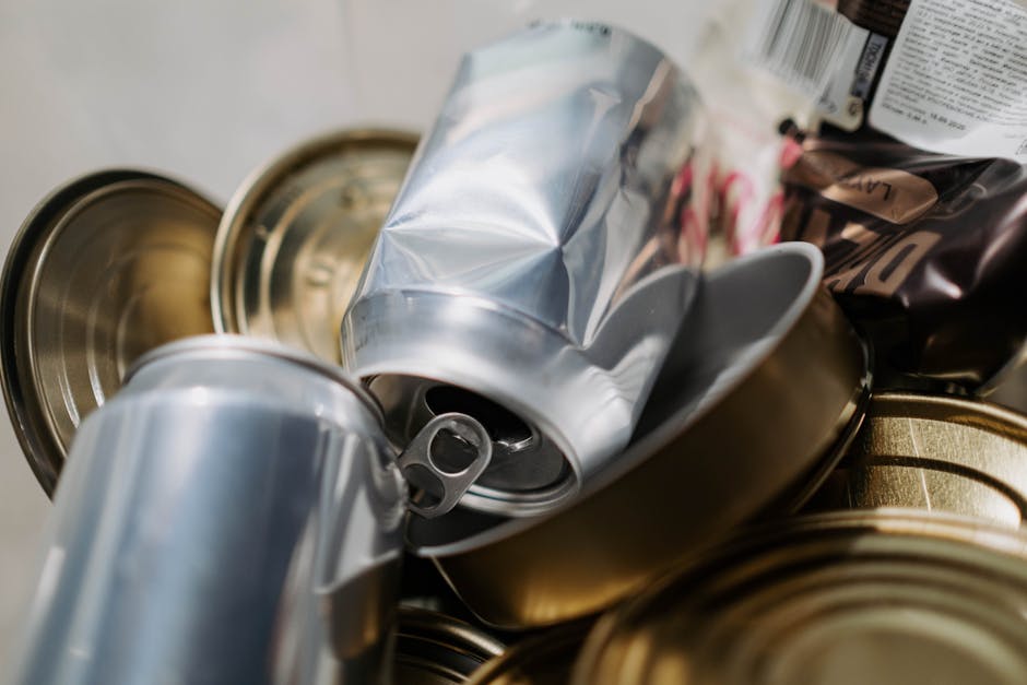 The image displays a close-up view of several crushed and partially flattened aluminium cans, with metallic silver surfaces reflecting light. The cans are stacked haphazardly, with some overlapping ea