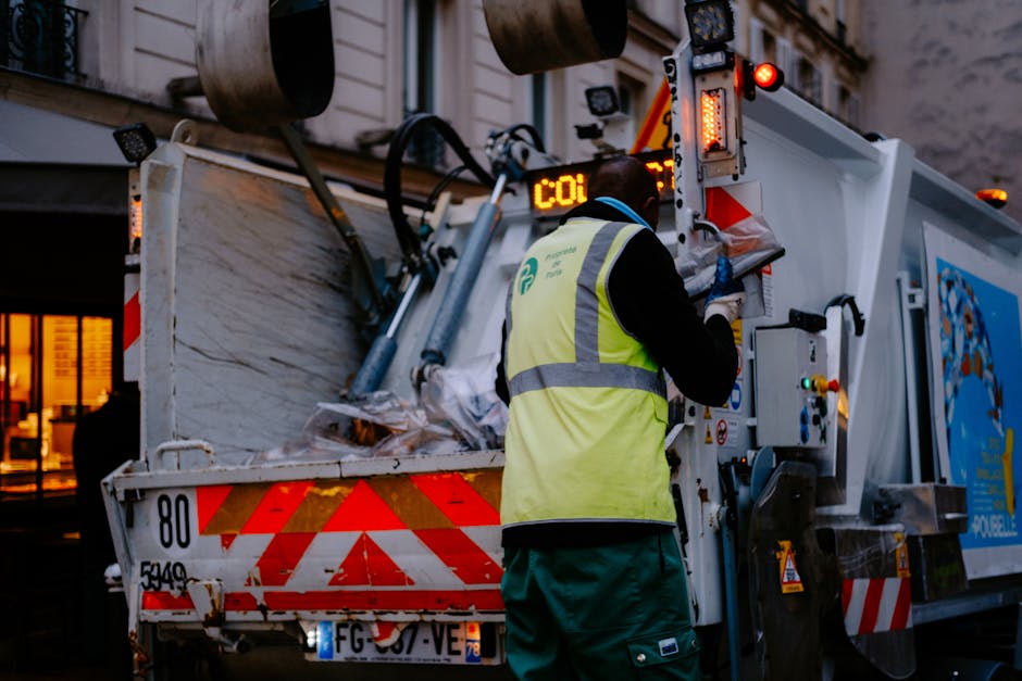 A waste collection worker, dressed in a black uniform with a high-visibility yellow vest featuring reflective grey stripes, is operating a compact waste collection vehicle on a city street during dusk