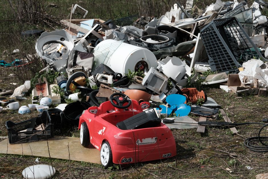 A large outdoor pile of mixed household waste and rubbish situated on a patch of bare earth and sparse grass, including broken electronic appliances such as a washing machine and miscellaneous metal a