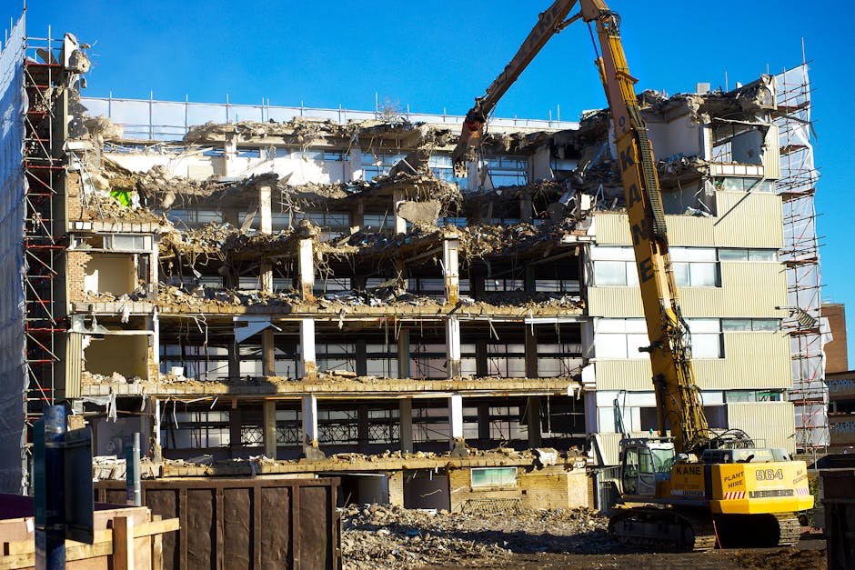 A multi-storey building under partial demolition, with several floors already torn down revealing exposed concrete, steel reinforcements, and remnants of interior walls. The structure includes section